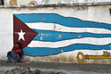 Un trabajador recostado a una pared en donde está pintada una bandera cubana Un trabajador recostado a una pared en donde está pintada una bandera cubana