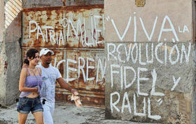 Una pareja de cubanos pasa junto a una puerta de metal oxidado y una pared deteriorada, con letreros de apoyo al régimen de los hermanos Castro