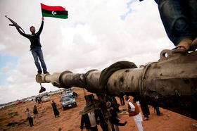 Un miliciano insurgente ondea la bandera de Libia subido encima de los restos de un carro blindado de las fuerzas de Muamar el Gadafi